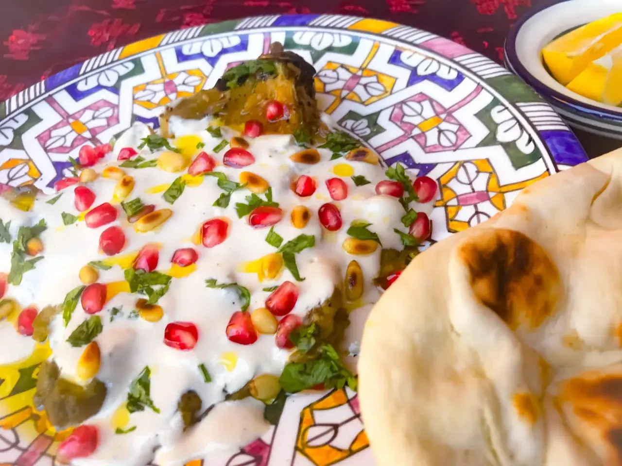 Un plat de mutabal crémeux, garni de grenades et de pignons, servi avec du pain pita sur une assiette décorée.