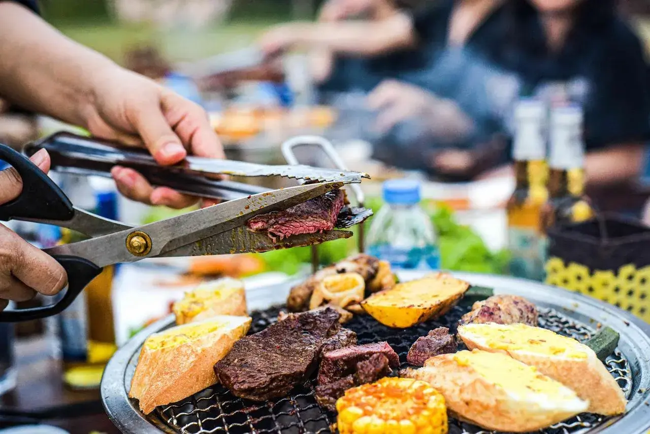 Une main coupe un steak juteux sur un gril lors d'une fête barbecue. Des pains grillés, des pommes de terre et du maïs complètent le festin.