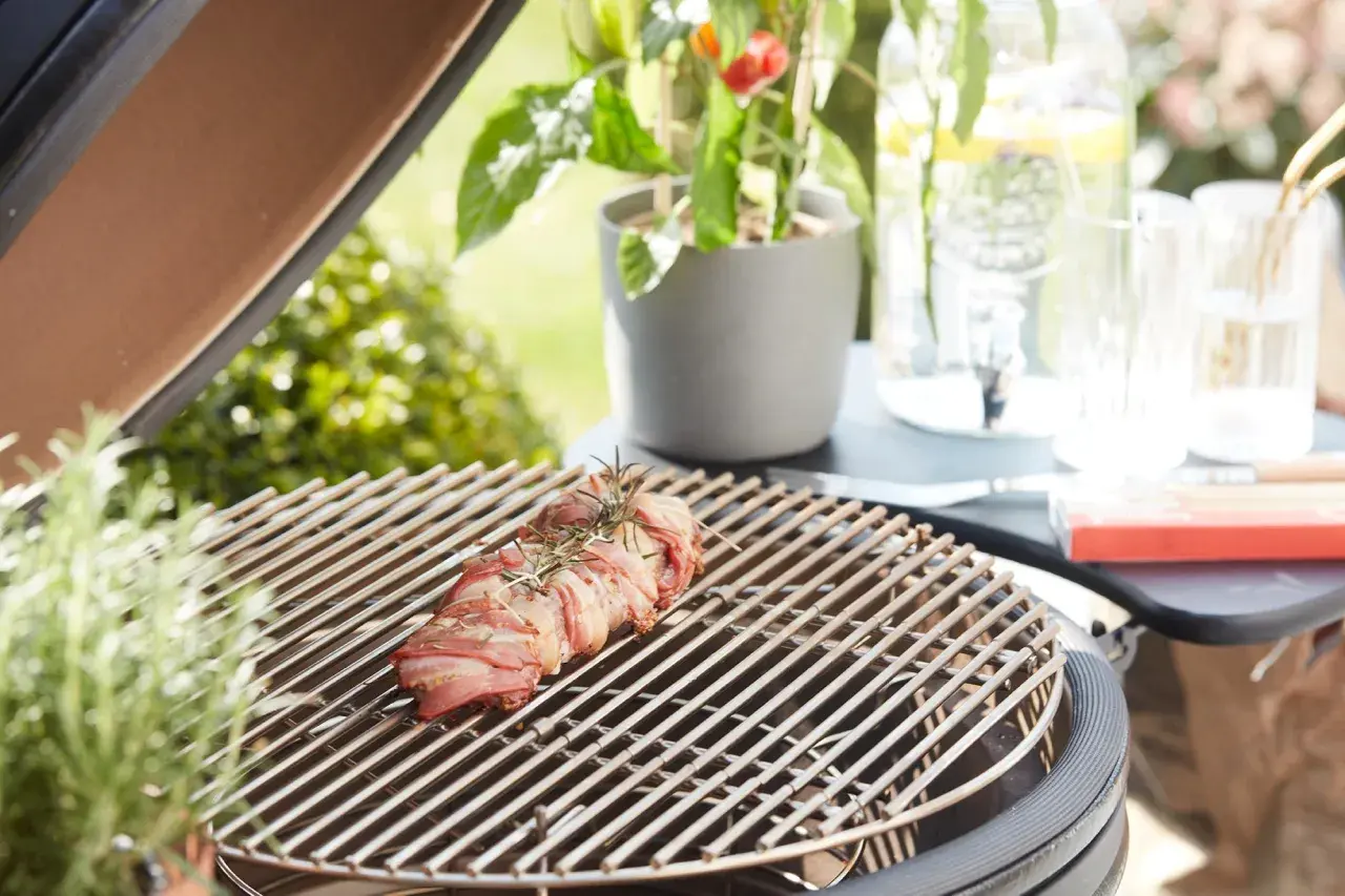Un rôti de viande enroulé de lard, prêt à griller. Une légère **fume bleu** s'échappe, promettant un repas savoureux en plein air.