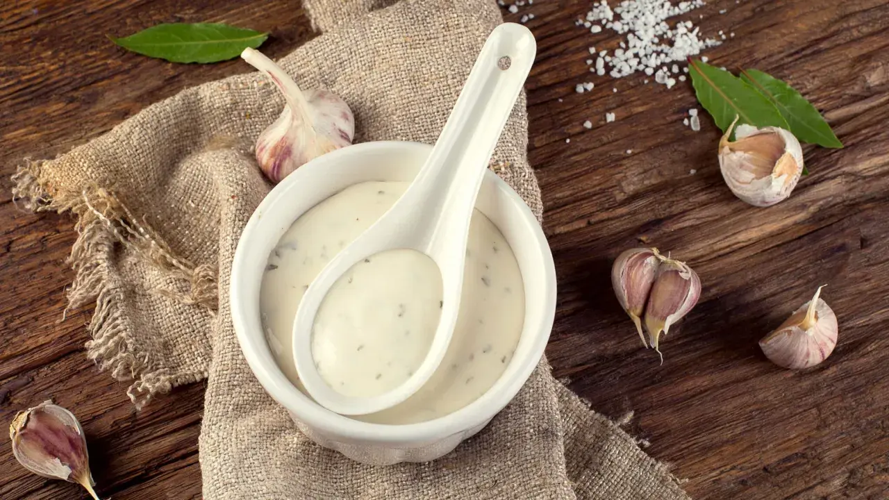 Sauce à l'ail crémeuse dans un bol blanc, avec une cuillère, des gousses d'ail et des feuilles de laurier sur une table en bois rustique.