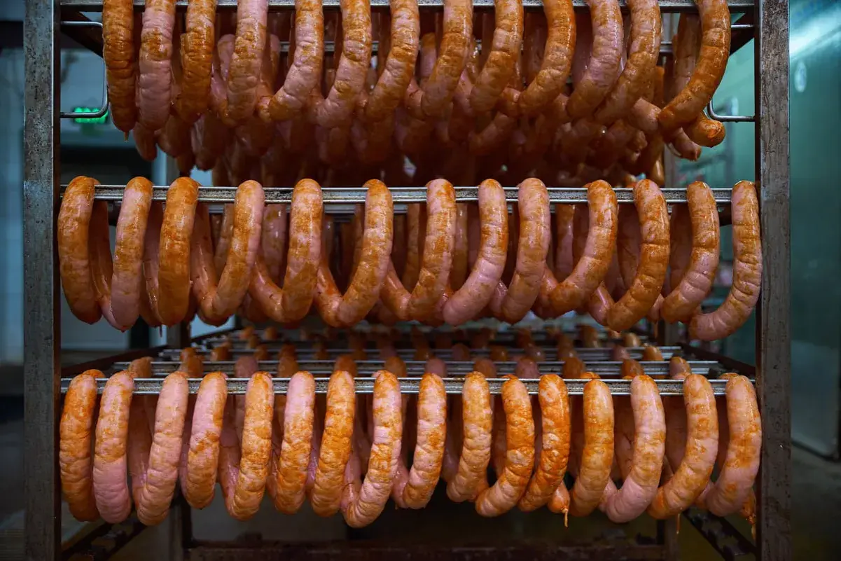 Des saucisses roses et orangées pendent en rangées sur des étagères métalliques, prêtes pour le processus de fumer des saucisses à froid.