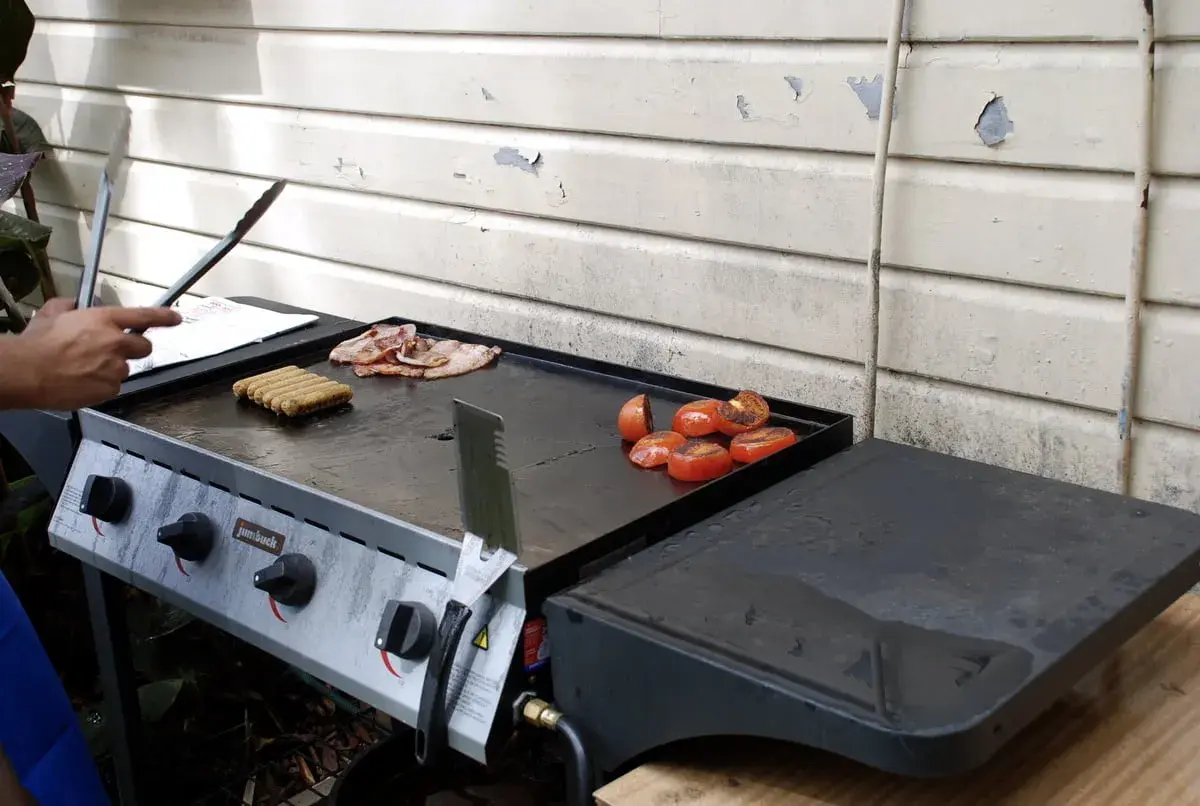 Comment allumer plancha gaz : des saucisses, du bacon et des tomates grillent sur la plaque.