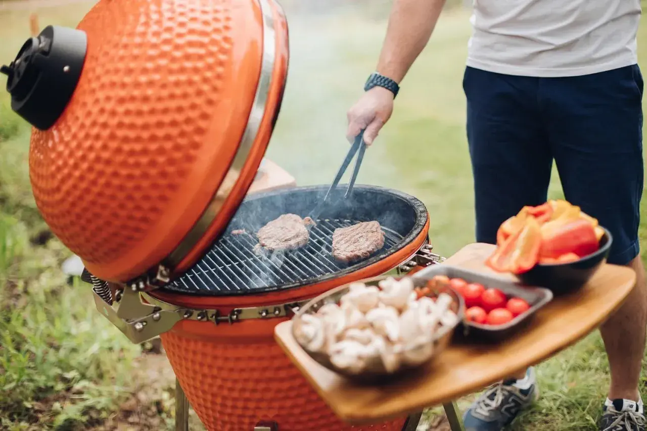 Un homme cuisine des steaks sur un kamado, c'est quoi ce barbecue en céramique orange. Des légumes sont prêts à être grillés.