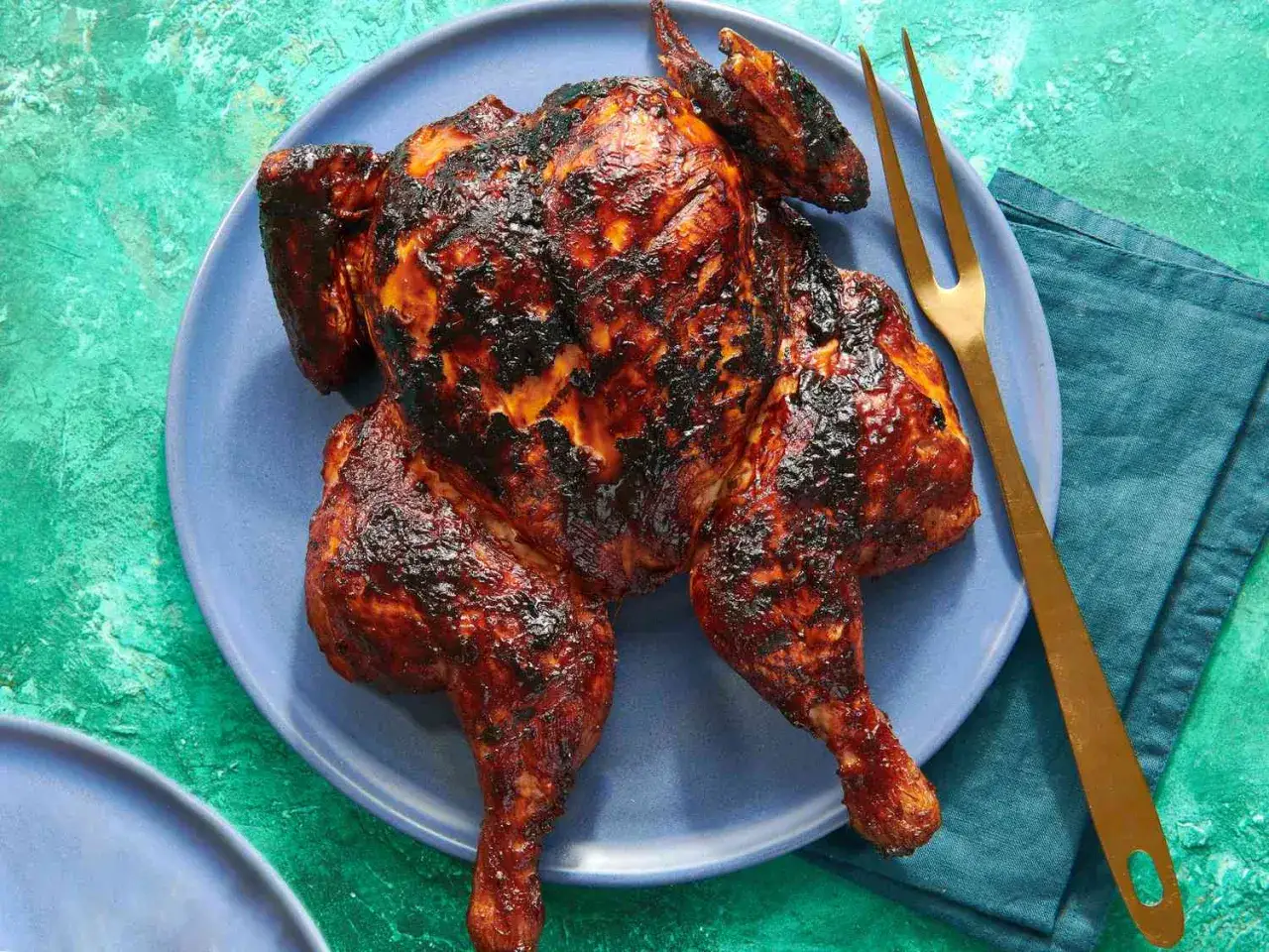 Un poulet entier au barbecue, la peau caram&eacute;lis&eacute;e et l&eacute;g&egrave;rement br&ucirc;l&eacute;e, est servi sur une assiette bleue avec une fourchette dor&eacute;e.