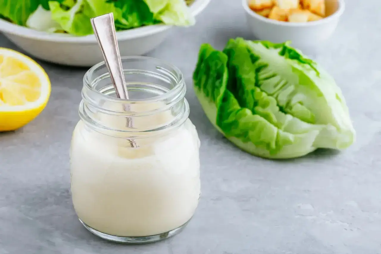Un pot de sauce césar crémeuse avec une cuillère, à côté d'une laitue romaine et d'un quartier de citron.