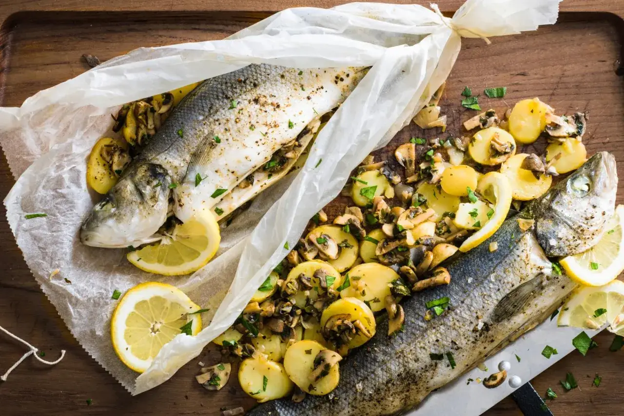 Deux bars en papillote au barbecue, garnis de pommes de terre et champignons, avec des rondelles de citron.