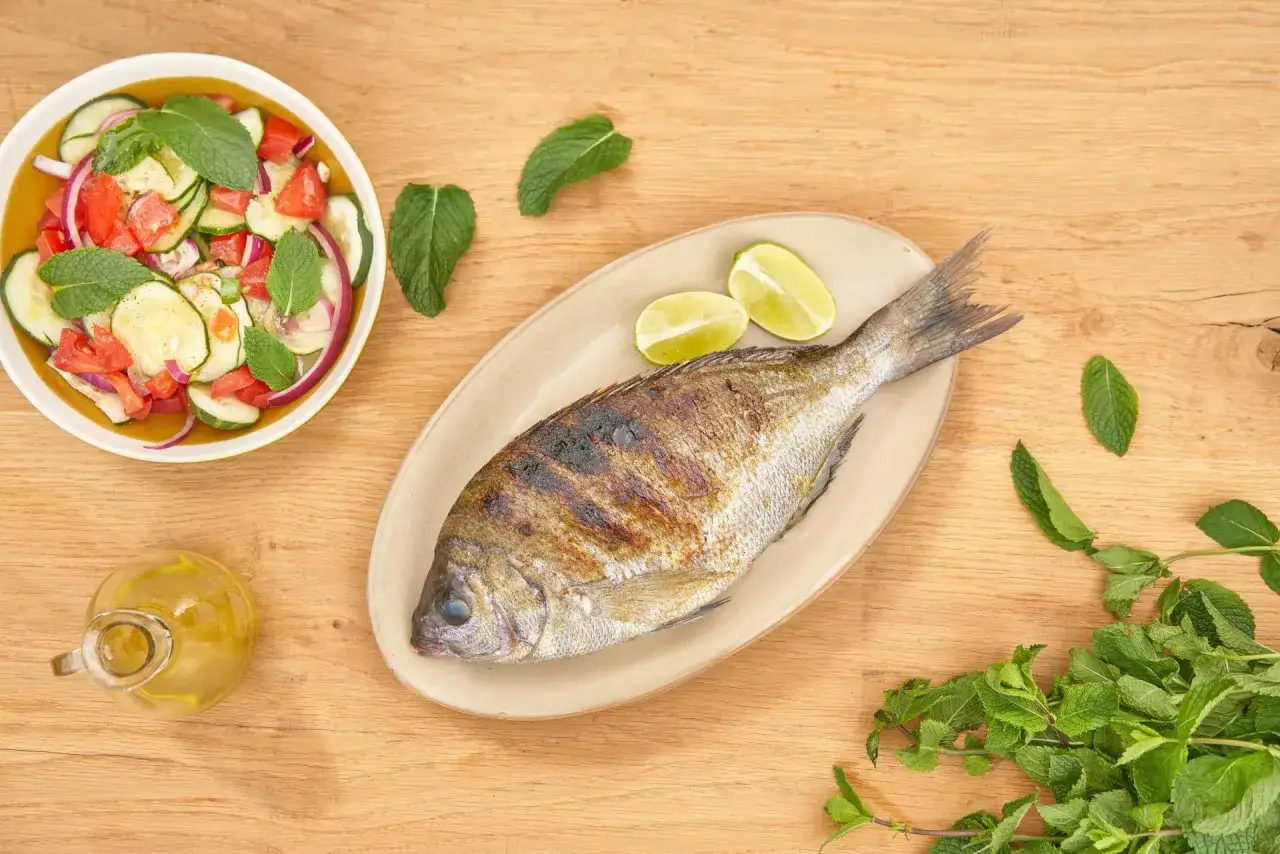Dorade grise grillée, prête pour la cuisson, accompagnée d'une salade fraîche et de quartiers de citron vert.