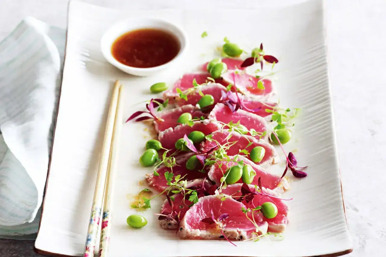 Tataki de thon finement tranché, garni de fèves edamame et de jeunes pousses, servi avec une sauce d'accompagnement.