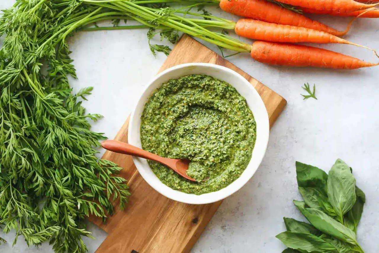 Un bol de pesto aux fanes de carottes, avec des carottes fraîches et du basilic à proximité.