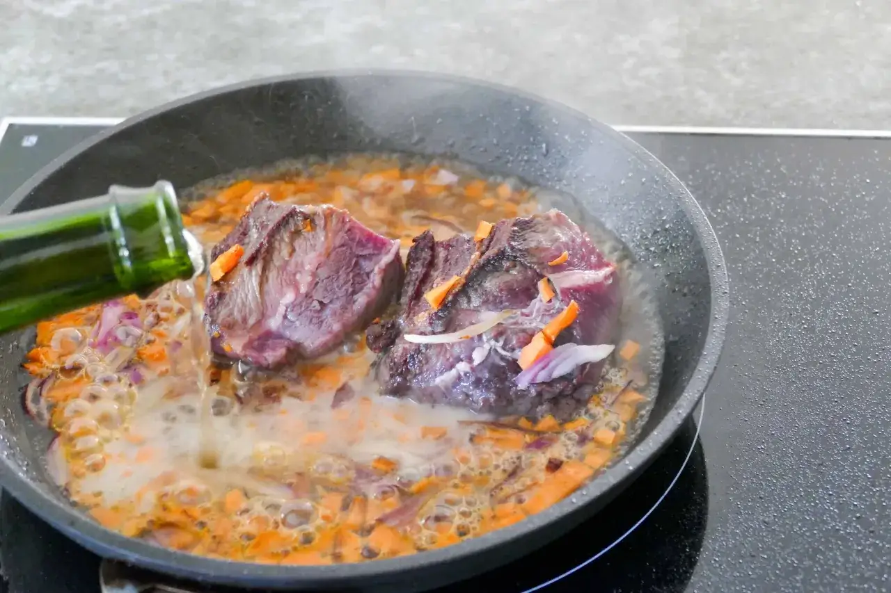 Deux morceaux de joue de b&oelig;uf mijotent dans une po&ecirc;le avec des carottes et des oignons. Du liquide est vers&eacute; d'une bouteille verte.