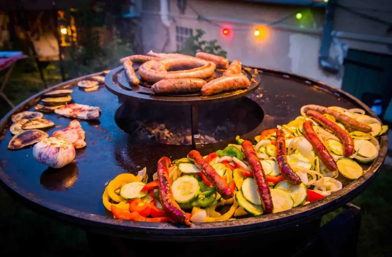 Oui, on peut faire des grillades sur un brasero ! Saucisses, légumes et viande cuisent sur cette grande plancha chauffée par le feu.