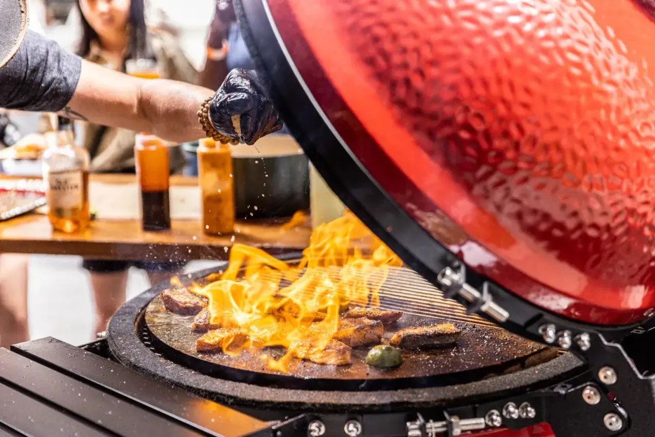 Un chef ganté ajoute une touche finale à la cuisson kamado. Des morceaux de viande flambent sur la grille, promettant un repas savoureux.