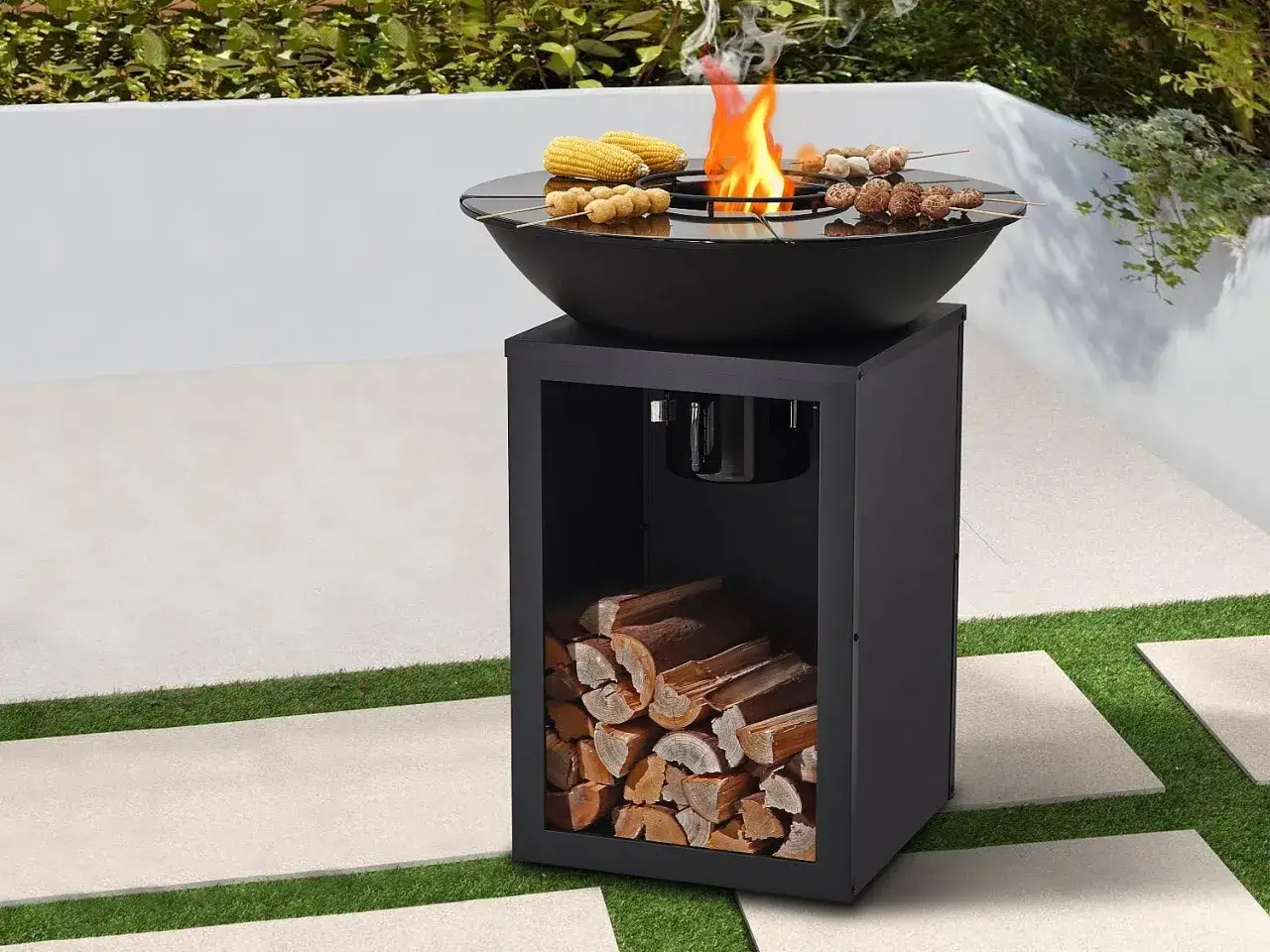Un brasero moderne avec du bois de chauffage rangé en dessous. Au-dessus, des brochettes et du maïs grillent sur des flammes vives.