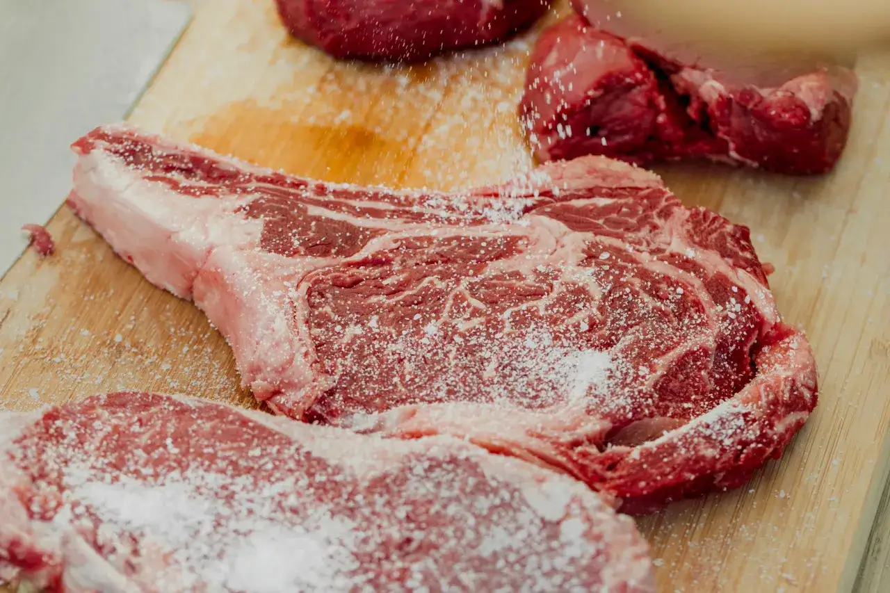 Gros morceaux de viande rouge, dont une côte de bœuf, sont assaisonnés de gros sel. Le persillage de la viande est bien visible, promettant une cuisson savoureuse.