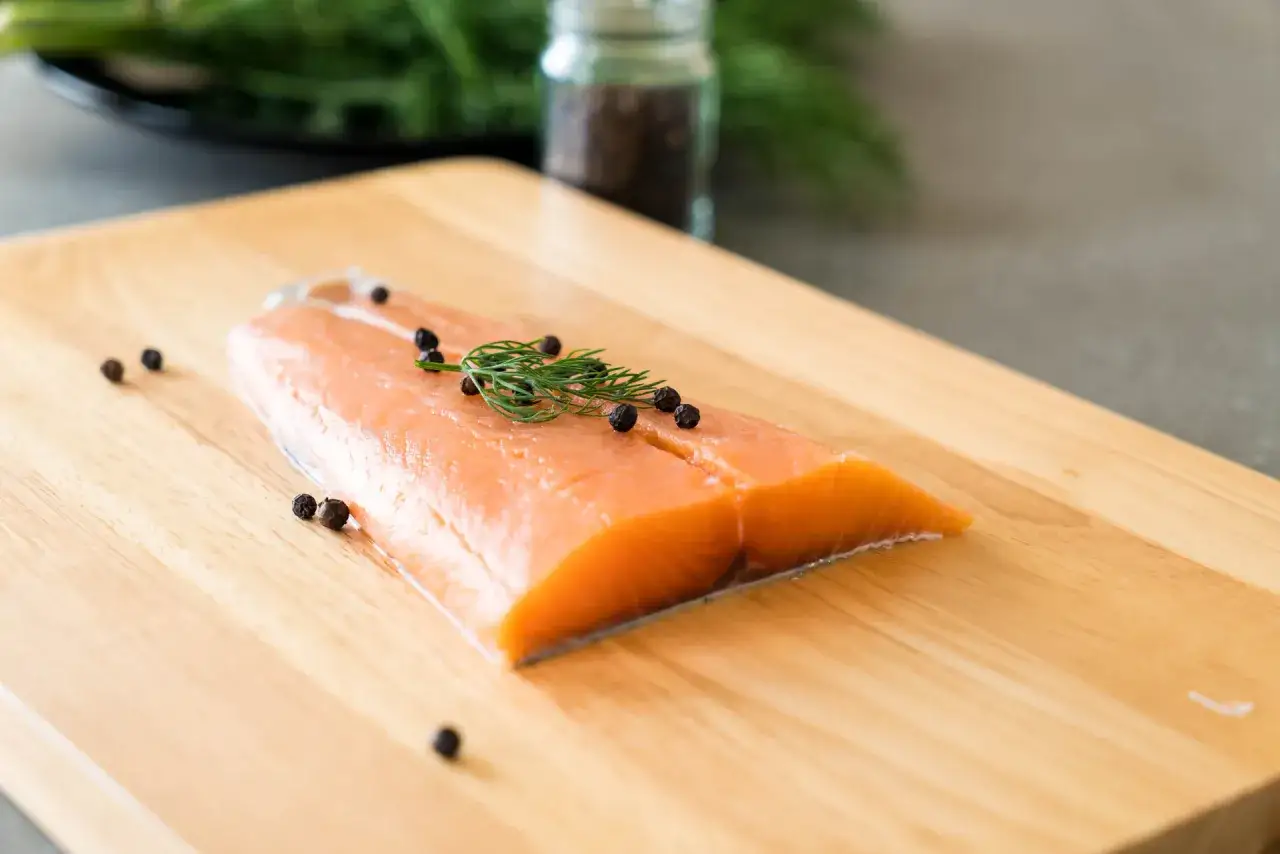 Filet de truite fumée prêt pour la cuisson, garni d'aneth et de grains de poivre noir sur une planche en bois.
