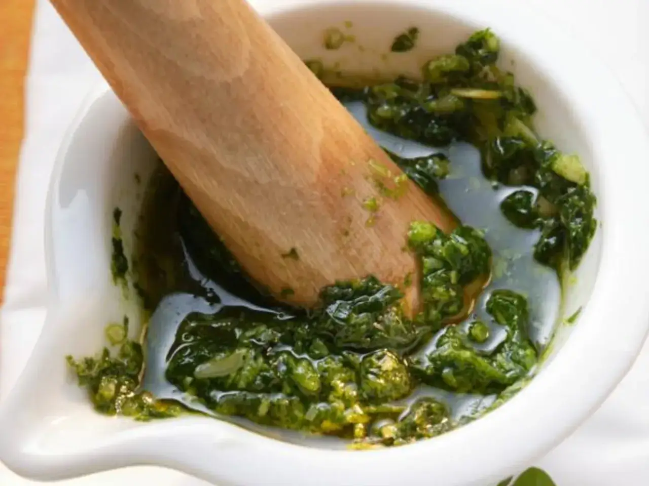 Un pilon en bois écrase des herbes fraîches dans un mortier blanc, créant un pistou parfumé.