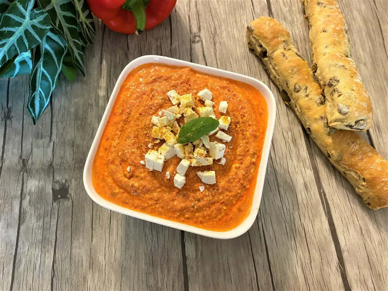 Un bol de poivronade, garni de dés de feta et d'une feuille de basilic, prêt à être dégusté avec des grissinis. Une recette estivale parfaite.