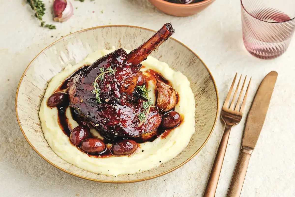 Une souris d'agneau confite, nappée d'une sauce brune et servie sur une purée onctueuse, accompagnée de quelques olives et d'ail rôti.