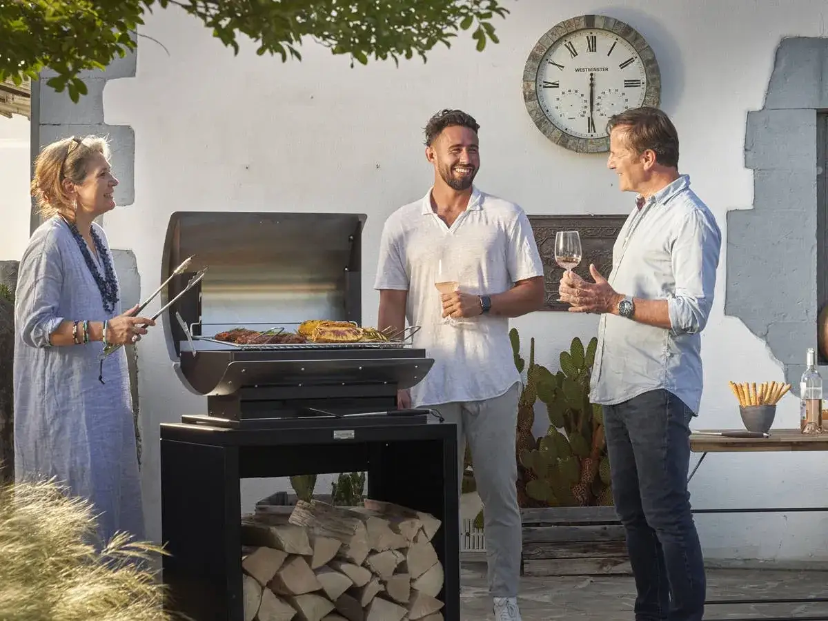 Une femme prépare une fete barbecue, tandis que deux hommes discutent avec des verres à la main.