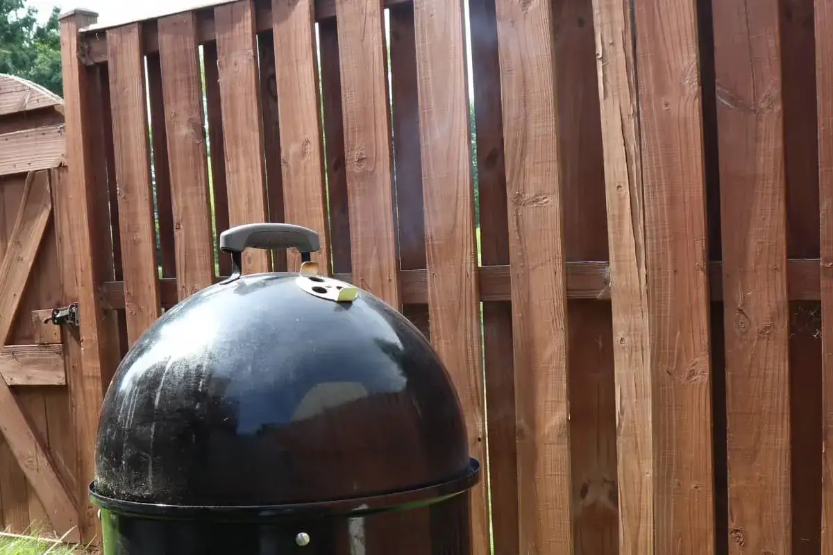 Un barbecue noir d&eacute;gage une fine fum&eacute;e bleu&acirc;tre devant une cl&ocirc;ture en bois.