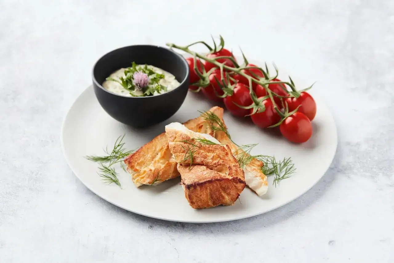Trois morceaux de dos de cabillaud fum&eacute;, servis avec une sauce cr&eacute;meuse, des tomates cerises et de l'aneth sur une assiette blanche.