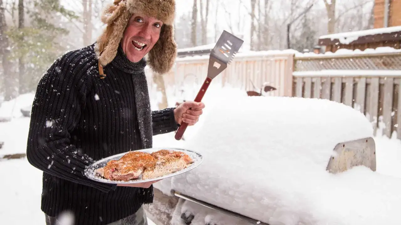 Un homme en bonnet de fourrure prépare un barbecue hivernal, tenant une assiette de viande assaisonnée et une spatule.
