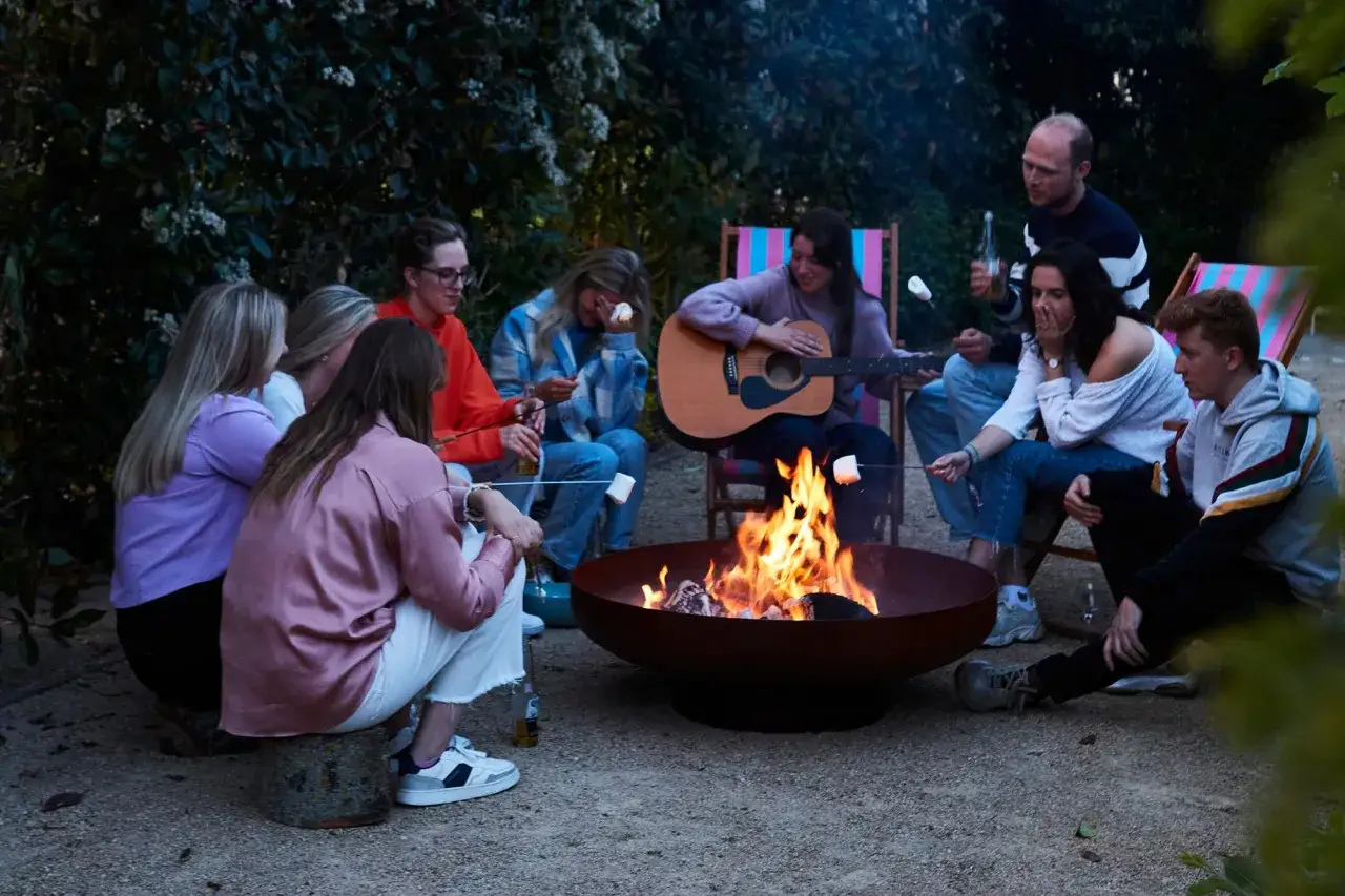 Une joyeuse bande d'amis profite d'une soiree brasero, grillant des guimauves autour d'un feu crépitant, tandis qu'une guitare accompagne l'ambiance.