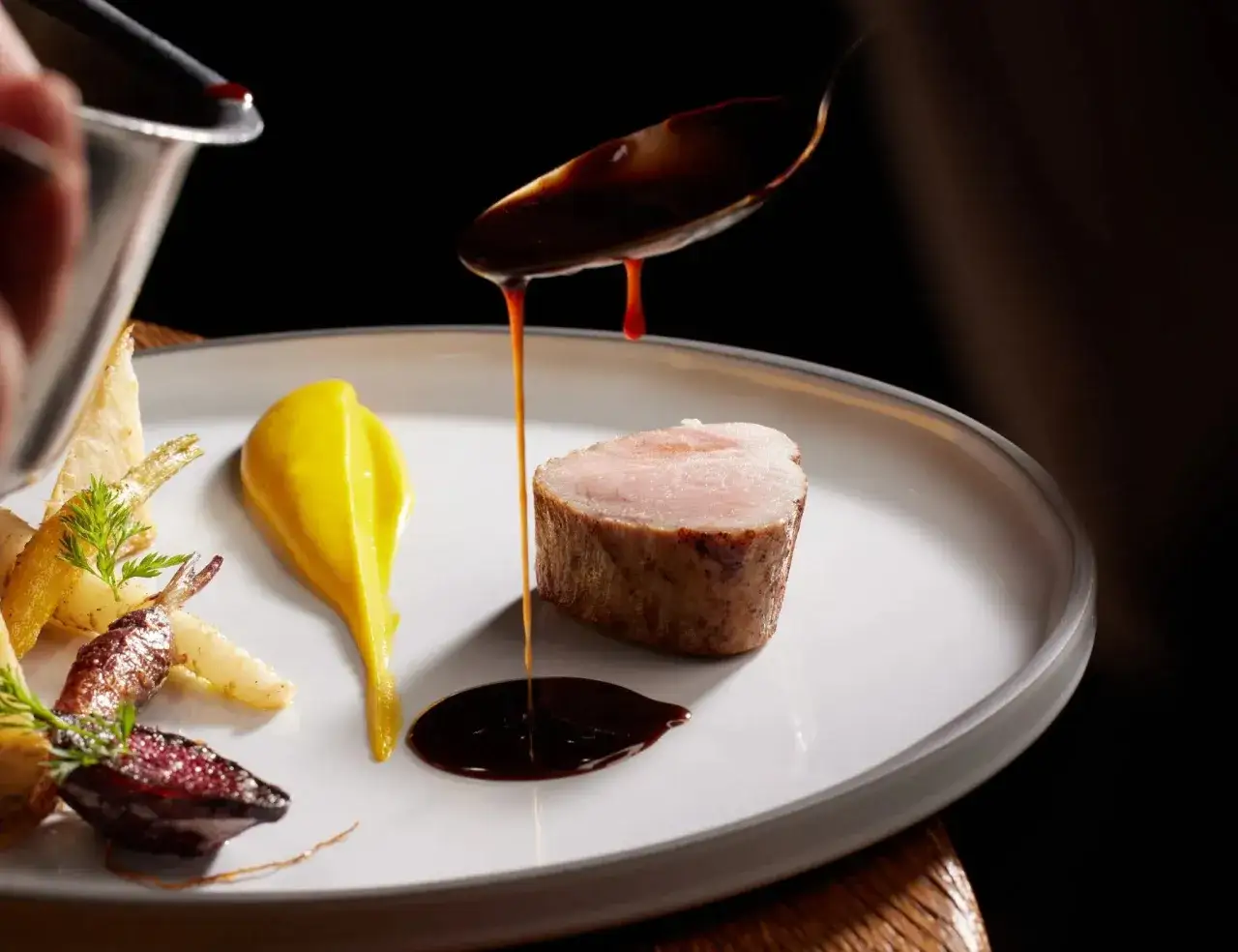 Une picanha de veau, sauce brune versée à la cuillère, purée jaune et légumes racines sur une assiette blanche.