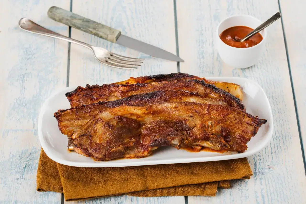 Trois morceaux de poitrine de porc barbecue, caramélisés et juteux, servis sur une assiette blanche avec une sauce barbecue.