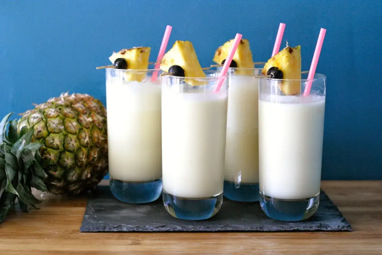 Quatre verres de pina colada, décorés de morceaux d'ananas et de cerises, avec un ananas entier à côté.