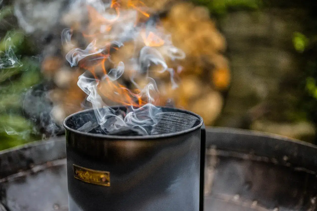 Le charbon s'embrase dans le cheminée, signe que le barbecue au charbon est prêt. La fumée blanche tourbillonne autour des flammes oranges.