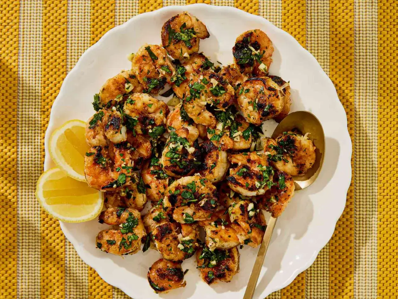 Brochette de scampis grillés, parsemés de persil frais et d'ail, servis avec des quartiers de citron sur une assiette blanche.